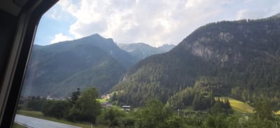 This media file captures a scenic view from a train window looking out at a mountain valley. The foreground is dominated by the dark frame of the train window and a grey road with white lines running diagonally across the bottom. Beyond the road, lush green trees and foliage cover the landscape.

In the middle ground, a small village with houses and a prominent church steeple with a red roof is nestled amongst the trees. The village appears peaceful and secluded. Further back, the scene is dominated by the imposing, tree-covered slopes of mountains that rise dramatically into the sky. The mountains are layered, with some peaks appearing rocky and others covered in dense forest. The sky above is a mix of blue with scattered white clouds, suggesting a partly sunny day.

The lighting indicates it is likely daytime, possibly late morning or early afternoon, with sunlight casting shadows and highlighting certain areas of the landscape, such as a bright green, sunlit hillside on the right. The overall mood is serene and picturesque, evoking a sense of travel and appreciation for natural beauty. There are no visible people or specific objects that stand out other than the natural elements and the man-made structures of the village. The location is identified as Schnann, Austria.