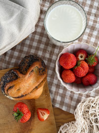 The image displays a still life of a breakfast setting on a brown and white checkered tablecloth. On a wooden cutting board, there is a twisted pastry sprinkled with poppy seeds and sugar. Beside the pastry, there is a whole strawberry and a halved strawberry. To the right of the cutting board, in a pink scalloped bowl, are several whole strawberries. A glass filled with white liquid, likely milk, is positioned behind the bowl of strawberries. In the upper left corner, a folded light-colored linen cloth partially covers a surface. A white mesh bag is visible in the lower right corner. The scene appears to be set indoors, with the lighting suggesting natural light coming from the side. There are no people present in the image. The setting does not provide specific clues to pinpoint a location within the city of Юг, Russia, as the elements are common to breakfast scenes worldwide.