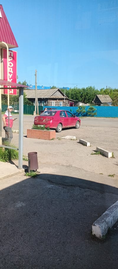 The image depicts a rural street scene in Киясово, Russia, under a clear blue sky. On the left, a red and white striped awning extends over a sidewalk, with a portion of a red sign that appears to read "ПРОДУК" (part of "ПРОДУКТЫ" meaning "groceries") visible. A person wearing a pink and white striped shirt, grey pants, and a blue cap is standing on the sidewalk, holding a smartphone. Adjacent to the sidewalk is a brown trash receptacle.
In the mid-ground, a red sedan is parked on a gravel and asphalt lot. The car has visible rust spots and appears to have stickers or decals on its rear. A brick planter box sits in front of the car. Behind the car, a bright blue fence runs along a property line. Beyond the fence, traditional wooden Russian houses with grey wooden roofs are visible, along with green trees and vegetation. A wooden utility pole stands in the background. The ground in the foreground and mid-ground is a mix of asphalt and gravel, with several white concrete parking stops present.