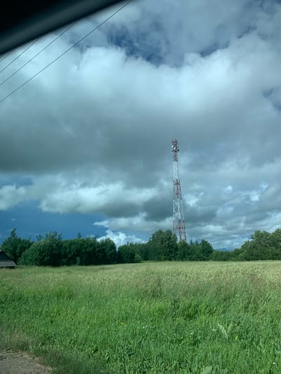 The image depicts a rural landscape under a dramatic, cloudy sky. In the center, a tall, red and white lattice telecommunications tower stands prominently against the dark, ominous clouds, which are lit by sunlight breaking through in some areas. In the foreground, a lush green field of tall grass stretches across the scene, with some scattered wildflowers and a small yellow flower visible. To the left of the field, a dark, rustic wooden structure, possibly a shed or barn, is partially visible behind a line of dense green trees. The trees form a dark line along the horizon, separating the field from the sky. Power lines are visible diagonally across the upper left corner of the image. The scene is likely located in a rural area such as Курапольскі сельскі Савет, Belarus, given the presence of agricultural land, a telecommunications tower, and a wooden structure. There are no people or animals visible in the image.