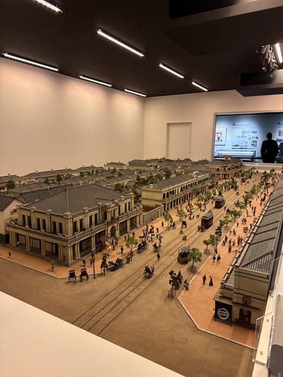 This image shows a detailed architectural model, likely a museum exhibit, depicting a historical street scene. The model features multi-story buildings with grey tiled roofs and intricate facades, suggesting an East Asian architectural style. Figures, carriages, and other miniature details populate the street, evoking a bustling historical atmosphere. Railway tracks run along the side of the street. In the background, a display screen shows historical images or information, and a person stands observing it. The scene is lit by museum-style lighting, highlighting the details of the model. The exhibit is possibly located in the city of Sumida, Japan.
