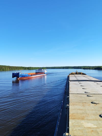 The image depicts a daytime scene on a wide river near Novodevich'e, Russia. The view is from the deck of a large cargo vessel, looking forward and to its port (left) side.

In the midground, another dry cargo ship, identified by the Cyrillic text "ОКСКИЙ-45М" (OKSKIY-45M) and "С-ПЕТЕРБУРГ" (S-PETERSBURG) on its white superstructure, is moving parallel to the camera vessel, heading from the left side of the frame towards the right. This vessel has a distinctive blue hull, a red bottom, and exposed orange-brown internal structures, indicating empty cargo holds. It leaves a prominent wake behind it.

The foreground shows the weathered, flat, grey steel deck of the camera vessel, featuring multiple oval-shaped access hatches. Blue railings and structural components run along the port side of this vessel. Towards the bow, yellow machinery elements are partially visible.

The river water is dark blue, marked with ripples and reflections. Both banks of the river are densely covered with green forests, extending to the horizon. The sky above is a clear, bright blue with no clouds, though a very faint contrail is discernible in the upper right corner. No people are visible in the image.
