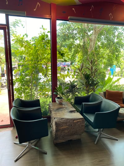 The image shows an indoor seating area in what appears to be a cafe or restaurant, with large windows overlooking lush greenery. The seating consists of four dark green, modern swivel chairs arranged around a rough-hewn wooden table. A small potted plant sits on the table. The chairs are positioned to face both the table and the expansive view outside.
The view through the windows reveals a vibrant outdoor scene. Dense foliage from various trees and plants fills the frame, suggesting a tropical environment. Sunlight filters through the leaves, creating a bright and airy atmosphere. In the distance, through the trees, glimpses of structures and possibly a street can be seen, hinting at the location being within a developed area. Decorative musical notes are painted on the red ceiling above the window. The floor is tiled in a wood-grain pattern. The overall impression is one of a comfortable and naturalistic indoor space connected to its tropical surroundings. Given the context provided about Bang Sare, Thailand, this scene likely depicts a cafe in that location, known for its natural beauty and relaxed atmosphere.
