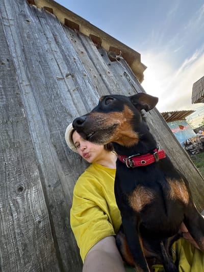 The image shows a person and a dog lying on a weathered wooden surface, likely a deck or porch. The dog, a medium-sized breed with black and tan markings, wears a red collar. The person is lying on their side, wearing a yellow shirt and a hat. The background includes a building with a partially visible mural and some greenery. The location is Усть-Тунтор, Russia. The composition is slightly tilted, giving the impression of a selfie taken from a low angle. The lighting suggests a sunny day.