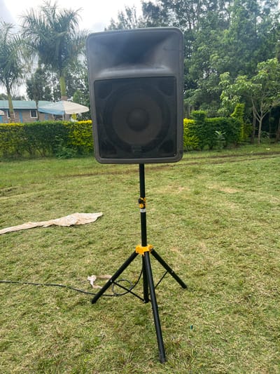An outdoor scene in Makuyu, Kenya, features a black PA speaker mounted on a tripod stand, positioned on a vibrant green grass lawn. The speaker, facing left in the image, has a protective metal grille covering its cone and horn, with a faint logo visible on the grille. The black tripod speaker stand has bright yellow accents at its adjustable joint. A black power or audio cable snakes across the grass from the base of the stand.
In the mid-ground and background, dense green foliage and tall trees create a natural setting. Among the trees, a few palm trees are distinctly visible, and a light-colored building with a roof is partially seen behind some hedges. A long, light-colored piece of fabric or paper is lying crumpled on the grass near the speaker stand.
The lighting suggests it is daytime, possibly with an overcast sky, as there are no strong shadows. The overall impression is that an audio system is being set up for an outdoor event or gathering in a garden or park-like environment.