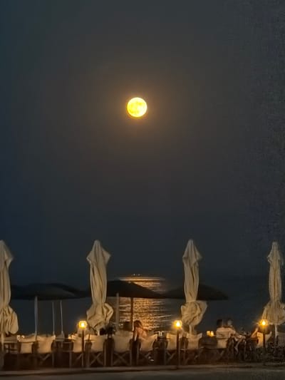 The image depicts a nighttime scene at a beachside restaurant in Nea Kallikratia, Greece. A bright, yellow full moon is prominently visible in the dark sky. In the foreground, several closed beach umbrellas stand upright, casting shadows. Tables and chairs are arranged in front of the umbrellas, suggesting a seating area for diners. Some tables are illuminated by small, decorative lamps. A few people are seated at the tables, with one person visible facing the camera, appearing to be at a table. The sea is visible in the background, with a distant boat or structure illuminated on the water. The overall atmosphere is serene and tranquil, with the moonlight creating a soft glow over the scene.