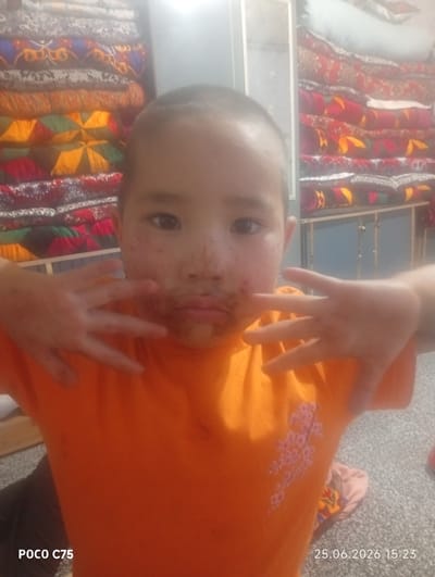 This media file features a young child in the foreground, looking directly at the camera with a playful expression. The child has reddish-brown marks around their mouth and on their face, possibly from food. They are wearing an orange t-shirt with a floral design on the chest. The child's hands are held up, with fingers spread, as if making a gesture.
In the background, there are stacks of colorful textiles, likely blankets or cushions, with intricate patterns. These are arranged on shelves or a rack. To the right, a light-colored object, possibly a pillow, is visible among the textiles. The overall setting appears to be indoors, with a slightly dim and warm lighting.
The timestamp "25.06.2026 15:23" is visible in the bottom right corner, indicating the date and time the photo was taken. The text "POCO C75" is present in the bottom left corner, likely indicating the device used to capture the image. The location context is Chon-Kara, Kyrgyzstan. No specific activity or event is clearly depicted, other than the child posing for the photo. The emotions conveyed are of a candid and perhaps mischievous moment.