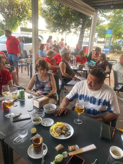 The image depicts an outdoor dining scene at a cafe or restaurant in San Pedro De Alcántara, Spain. Several people are seated at black tables, some of whom appear to be engaged in conversation and enjoying drinks and snacks.

In the foreground, a man with a tie-dye t-shirt is seated at a table, looking to his right, with a glass of beer in front of him. To his left, a woman wearing a sleeveless top with a printed image is also seated at a table, looking down and touching a small wooden box. A glass of beer, a can of soda, and a plate of snacks are on the table in front of her. Further to the left, a man in a red polo shirt and jeans is standing and looking to his left.

In the background, other patrons are visible, including women in casual attire and a man in a white shirt. A car and a public telephone booth are visible in the distance, suggesting an urban or semi-urban setting. The overall atmosphere appears relaxed and social, with ample natural light indicating it is daytime. Trees and foliage are present, contributing to the outdoor ambiance.