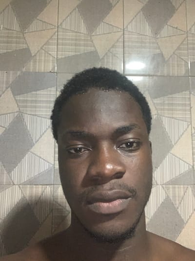 A dark-skinned man with short, dark hair and a trimmed beard faces the camera. His expression is neutral. He has a broad nose, full lips, and dark eyes. The background features patterned gray and beige floor tiles with geometric designs. The angle of the image is slightly tilted. A small area of bare skin is visible in the upper right corner.