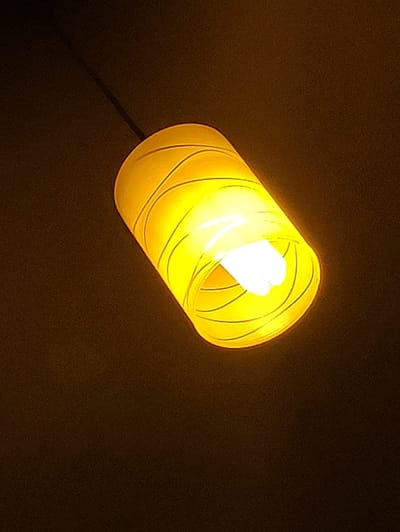 A hanging light fixture with a yellow, cylindrical shade illuminates a dark, textured background. The shade has a coiled pattern created by darker lines against the bright yellow material. Inside the shade, a white light bulb is visible, emitting a strong glow that causes the shade to appear translucent. The light fixture is suspended from a dark cord, which extends upwards and out of frame. The overall impression is one of artificial light in a dimly lit, indoor space. The image does not contain any information about the city of Krushev Dol, Bulgaria.