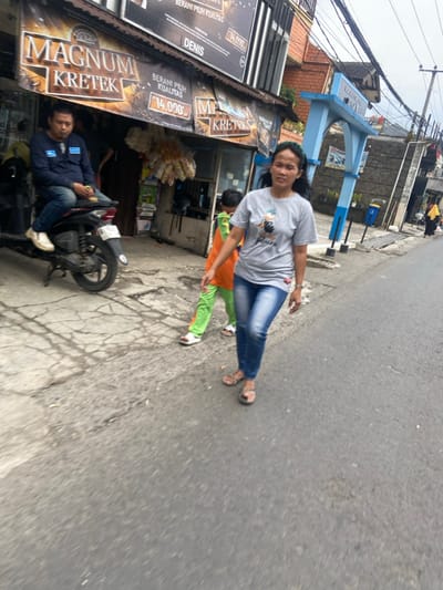 The scene is a street in Kecamatan Cimahi Selatan, Indonesia. A woman in a grey t-shirt and blue jeans is walking down the middle of the street with a young child in a green and orange tracksuit. The child is slightly behind the woman. The woman is looking straight ahead, and the child is looking to the left. The woman is wearing sandals, and the child is wearing white sneakers. The woman is walking on the left side of the street, and the child is walking on the right side of the street. There is a motorcycle parked on the right side of the street, and a man sitting on it. The man is wearing a dark blue shirt and black pants. He is looking straight ahead. There is a blue archway structure on the right side of the street. There are some buildings in the background, and there are power lines running across the street. The road is paved and has a few cracks. The sidewalk on the left side of the street is made of cobblestones. The sidewalk on the right side of the street is made of concrete. There are a few trees and bushes on the left side of the street. There are some trash cans on the right side of the street. There are some signs and banners on the left side of the street. The scene is taken in the daytime. The weather is clear.