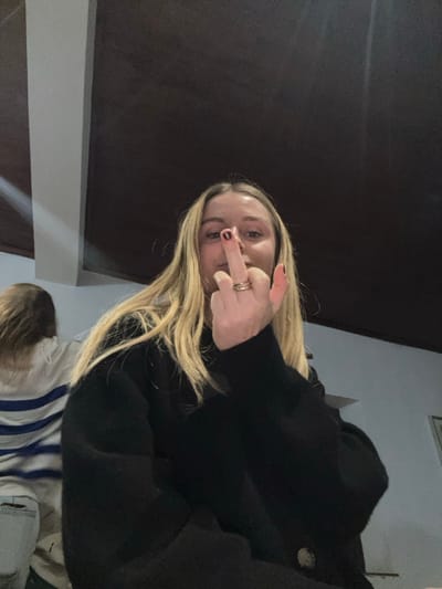 A woman with long blonde hair and a black cardigan is giving the middle finger. She has dark red nail polish and a ring on her middle finger. The background is dark, possibly a ceiling. In the bottom corner, there is a small child with light hair and a striped shirt. The picture is taken from a high angle, looking down on the subjects.