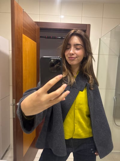 A female individual is depicted taking a mirror selfie indoors in Nogueira e Silva Escura, Portugal. The subject, with light brown hair and light complexion, smiles with closed eyes, holding a dark-colored smartphone in her right hand, angled to capture her reflection. She is dressed in a bright yellow long-sleeved top beneath a dark grey, textured, open-front cape or poncho, and dark blue jeans. Her fingernails are painted a light, neutral color. The environment features white rectangular tiled walls. A partially open wooden door with a horizontal two-tone design and a metallic handle is visible on the left. Above the door frame, a black and red pull-up bar is installed. A transparent glass panel stands to the right.