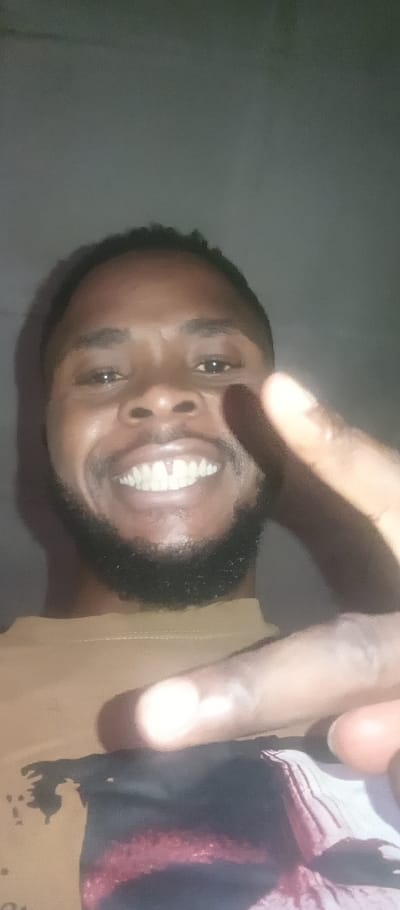 A smiling Black man with a beard poses for a selfie. He’s wearing a tan t-shirt with a colorful graphic design at the bottom. He’s giving a peace sign with his right hand, obscuring part of his face. The lighting is poor, and the background is a plain, dark gray. The focus is on the man's face, particularly his smile and white teeth.