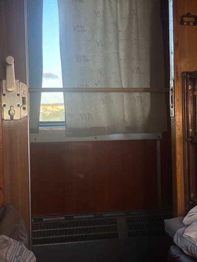 A close-up shot of a train window covered by a slightly raised, patterned curtain. A wooden rod is positioned vertically in front of the curtain. Below the window, a metal latch mechanism is visible. To the right of the window, there's a dark brown, possibly wooden panel and a gray, ventilated section. Above, there's dark brown wooden panelling with some metal fixtures and some light coloured fabric/clothes.