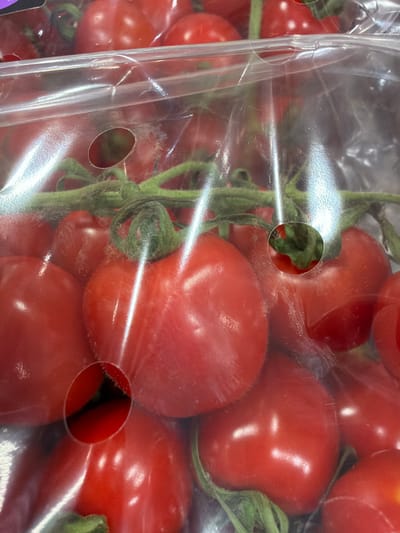 The image shows a close-up of a package of ripe, red cherry tomatoes. The tomatoes are attached to green stems and are partially obscured by the clear plastic packaging. There are small, circular holes in the plastic packaging, allowing for some visibility of the tomatoes within. The lighting is bright, casting reflections on the plastic and the tomatoes. The scene appears to be a retail environment. The location of Krustkalni, Latvia is not depicted or implied in the image.