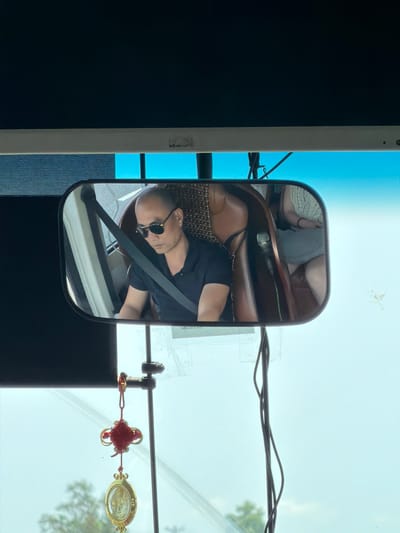 The reflection in a bus side mirror shows a person wearing sunglasses and a seatbelt. A red Chinese knot and a religious pendant hang from the bus's interior near the windshield. Outside the bus, power lines are visible against a hazy sky and some tree branches. The location is likely in Hải Phòng, Vietnam, based on the red Chinese knot and religious pendant which are commonly found in this area of the world.