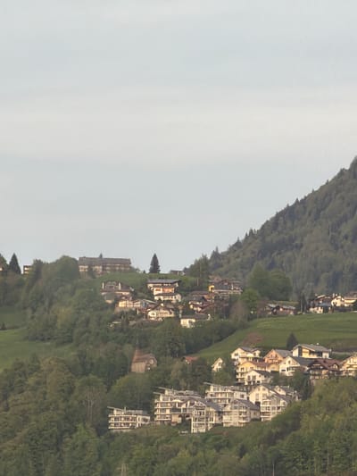This image presents a scenic view of a village nestled along a verdant hillside, under a soft, diffused sky. The primary subjects are the natural landscape and human habitation.
The scene is dominated by a tree-covered mountain or steep hill, which occupies the upper left portion of the frame and slopes gently downwards towards the middle and right. This hillside is densely covered with lush green trees and other vegetation, indicating a rich natural environment. Below the main slope, and extending across flatter terrain to the right and bottom of the image, is a concentrated settlement.
Numerous buildings, predominantly houses and apartment blocks, are constructed along the hillside and on the adjacent flat ground. Many of these structures are multi-story, featuring white or light-colored facades, often with numerous windows and balconies. The architectural styles appear varied but cohesive, characteristic of a European mountain or lakeside village. Some buildings are quite large, suggesting multi-unit dwellings. Green grassy areas and scattered trees are interspersed among the buildings, blending the built environment with nature.
The upper left section of the image is filled with a bright, overcast sky. The absence of harsh shadows or direct sunlight suggests either an overcast day, or perhaps early morning or late afternoon light. No specific activities or people are visible within the frame, portraying a serene and quiet atmosphere. There is no discernible text in the image. The appearance of the village integrated into the mountainous landscape strongly aligns with the provided location context of Beckenried, Switzerland.