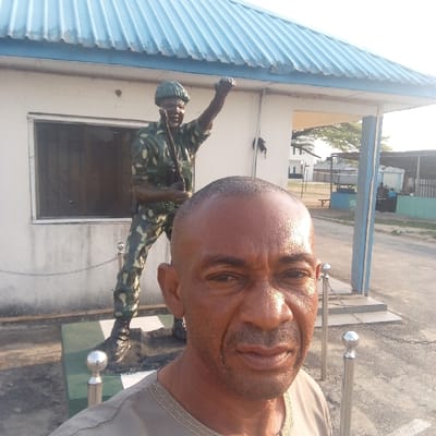 An outdoor daylight photograph in Koko, Nigeria, features an adult male positioned for a selfie in the foreground, his upper torso visible. He has a shaved head, dark skin, and a light goatee, wearing a grey shirt with patterned trim, and looks directly at the camera. Behind him, to the left, stands a life-sized statue of a soldier, painted in green and brown camouflage, holding a rifle in the right hand and with the left fist raised. The statue rests on a rectangular green and white pedestal. Immediately behind the statue is a single-story building with white walls, a barred window, and a light blue corrugated metal roof. To the right, a series of silver bollards connected by chains mark a boundary. The background shows an open area with visible ground, distant trees, and additional structures under a bright sky.