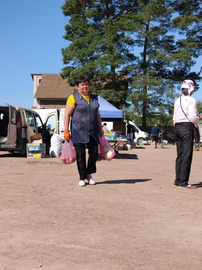 A woman in a blue polka-dot vest over a yellow t-shirt and black pants walks with two pink plastic bags filled with red onions in a dirt lot. She wears orange gloves and white shoes. Behind her, a light-colored van with its rear doors open is parked next to a white cargo van. In the background, a building with a brown-tiled roof stands under tall green trees, and a blue tent is set up. Another woman in a pink shirt and black pants stands facing away from the camera to the right, with a dark bag slung over her shoulder. The scene appears to be a market or an outdoor gathering in Irshansk, Ukraine, under a clear blue sky.