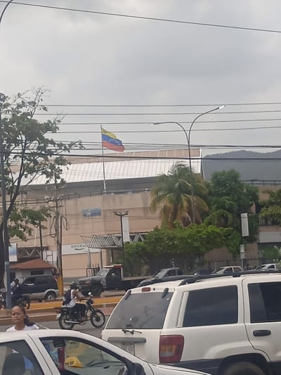The image captures a street scene in Puerto La Cruz, Venezuela, under an overcast sky. In the background, a large building with a distinctive sloped roof is visible, adorned with a Venezuelan flag flying from a pole on its upper level. The building appears to be a commercial establishment, with signage that includes "BANCO PLAZA" visible.

In the foreground, several vehicles are present. A white SUV, possibly a Jeep Grand Cherokee, is parked in the right foreground. To its left, a motorcycle carries a rider wearing a backpack and a helmet, navigating the street. Further left, a black SUV is parked.

A woman in a white shirt is walking on the sidewalk, looking towards the viewer. A portion of a white car is visible in the very bottom left of the frame. The street itself has yellow and white lane markings, indicating a traffic intersection. Power lines crisscross the sky, and lush green trees and palm trees are scattered throughout the scene, suggesting a tropical or subtropical climate. The overall impression is of a busy urban environment in daylight.