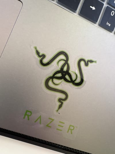 This close-up, high-angle shot shows a silver laptop with a Razer logo sticker and some keyboard keys visible. The sticker is a stylized black snake emblem with a green outline, placed on the laptop's lid. To the right of the snake logo, the word "RAZER" is visible in green letters on a transparent sticker, which is partially covering the lid. Some keys of the laptop's keyboard are also visible on the left side of the image, including an upward-pointing arrow key and a key labeled "0" and "Ins". The setting appears to be indoors, with neutral lighting. There are no people or specific environmental details that suggest a time of day, weather, or a particular location beyond what can be inferred from the object itself. The overall impression is of a close-up detail of a gaming laptop.