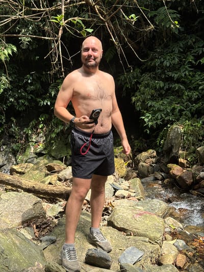 A bald man in black shorts and grey sneakers stands among rocks and foliage, holding a black device with a red cord in his hand. He is shirtless and appears to be in an outdoor environment near a stream or waterfall. The backdrop features lush green vegetation and rocky terrain, suggesting a natural setting. The image was taken in Suối Giàng, Vietnam.