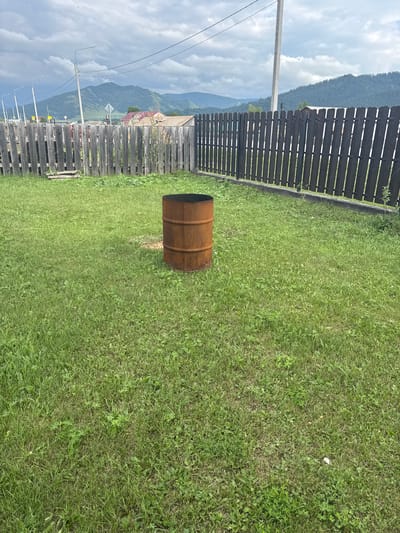 An exterior, medium shot captures a rustic backyard in Yabogan, Russia, under a cloudy sky. In the foreground, a weathered, rusty metal barrel sits prominently on a patch of green grass. The barrel appears to be a former industrial drum, showing signs of age and exposure to the elements.

Behind the barrel, a wooden fence stretches across the frame, dividing the yard from what lies beyond. The fence is constructed from vertical wooden planks, some of which are visibly aged and weathered. Beyond the fence, more rustic wooden structures and buildings with red roofs are visible. In the distance, rolling green hills and mountains are shrouded in a light haze, suggesting a rural or mountainous landscape. The sky is filled with heavy, grey clouds, hinting at the possibility of rain. The overall scene conveys a sense of simplicity and a connection to nature, typical of rural Russian settings. There are no people or animals present in the frame.