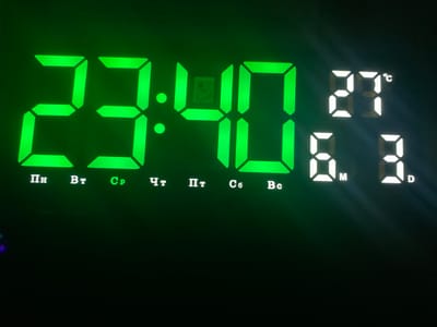 This media file captures a close-up of a digital display, likely an alarm or wall clock, set in an indoor environment in Verkhnevesyoloye, Russia. The background is dark, suggesting nighttime or a dimly lit room.

The most prominent feature is the time, displayed in bright green as "23:40", indicating 11:40 PM. To the right of the time, the temperature is shown as "27°C" in a dimmer white illumination. Below the temperature, the date "6.3." is visible, presumably March 6th, accompanied by small "M" (Month) and "D" (Day) indicators.

Beneath the main time display, a row of Cyrillic abbreviations for the days of the week is presented: "Пн" (Monday), "Вт" (Tuesday), "Ср" (Wednesday), "Чт" (Thursday), "Пт" (Friday), "Сб" (Saturday), and "Вс" (Sunday). Among these, "Ср" (Wednesday) is distinctly lit in green, signifying the current day. The other day abbreviations are unlit or very faint. An indistinct, small rectangular component or sensor is noticeable within the segment of the '0' in the minutes display. The image focuses entirely on the glowing display, providing functional information about the current time, temperature, and date.
