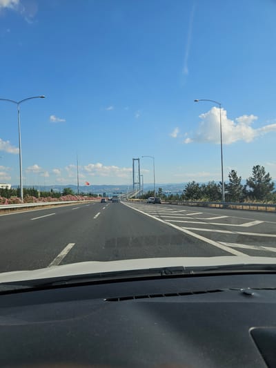 The image shows a view from a vehicle driving on a multi-lane highway. The highway has clear lane markings and a guardrail on the right side. In the distance, a suspension bridge is visible, with its towers and cables stretching across the frame. The sky is blue with scattered white clouds. The surrounding landscape includes trees and some buildings. A Turkish flag is flying on a pole along the side of the road. The scene appears to be in Dilovası, Turkey, possibly on the approach to the Osman Gazi Bridge.