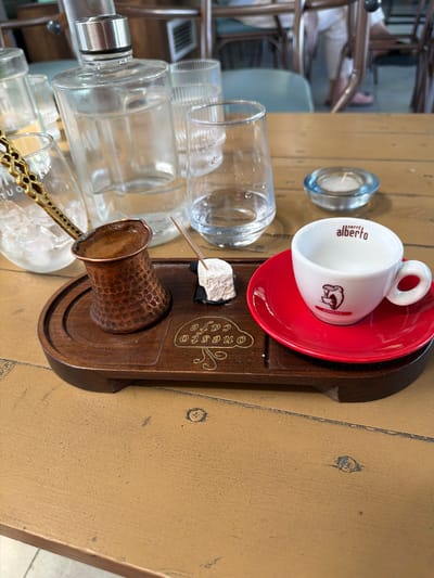 A close-up, high-angle shot shows a wooden tray with a small copper pot filled with coffee, a cube of Turkish delight, a white espresso cup on a red saucer, and a glass of water. A bottle of water, other glasses, and a small candle in a blue glass holder are also visible on a wooden table. The setting appears to be outdoors, possibly at an establishment named "onesto cafe" in Drama, Greece, based on the embossed text on the tray. The overall scene evokes a relaxed, cafe atmosphere.
