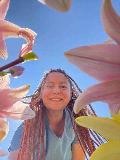 A low-angle shot captures a smiling woman with long, braided hair, possibly dreadlocks, in Tatarstan, Russia. She is looking up at the camera, framed by large, soft pink and yellow lily petals. The background is a clear blue sky. The woman is wearing a light blue t-shirt. The lighting suggests it is daytime, likely outdoors. The composition is dynamic, with the flowers creating a natural frame around the woman's face.