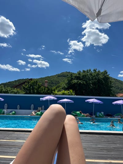 In Varvara, Bulgaria, on a bright, sunny day, a person's legs are in the foreground, lying on a wooden deck. Beyond the deck, a swimming pool filled with clear blue water is visible. Several people are in the pool, some swimming and others relaxing. Along the poolside, there are lounge chairs with purple umbrellas providing shade. In the background, a lush green hillside with trees and a house can be seen under a sky with white, fluffy clouds.