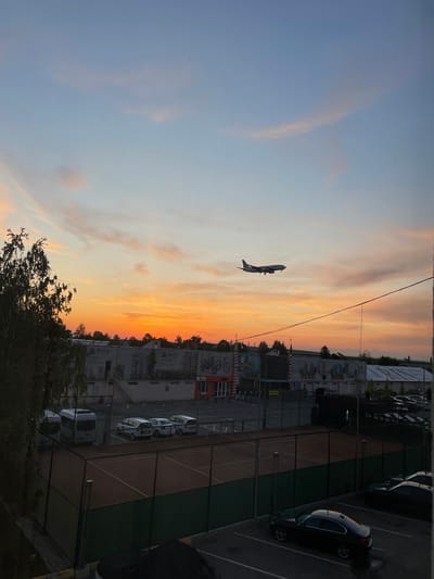 The original image, rotated 90 degrees clockwise, captures a vibrant golden hour scene in Otopeni, Romania, likely in the vicinity of Henri Coandă International Airport. The sky dominates the upper portion, painted with warm gradients of orange, yellow, and soft blue, indicative of either sunset or sunrise. Thin, wispy clouds are scattered across the expansive sky.

A commercial airplane, silhouetted against the bright backdrop, is a central subject, appearing in the upper-left (when correctly oriented). Its low altitude suggests it is either on approach for landing or has recently taken off. Stretching across the sky, faint power lines or utility cables are visible.

Below the colorful sky, the ground level presents an urban or suburban landscape. To the right, several multi-story buildings line the horizon. Some of these structures appear to be under construction or feature large advertisements, with the text "HILS" clearly visible on at least two buildings, along with a partial phone number "07...74". A red and white striped barrier or pole is also noticeable. Adjacent to these buildings is a sports court, likely a tennis or padel court, characterized by its distinct green and reddish-brown sections and surrounding fencing. Various parked vehicles, including a dark sedan and several lighter-colored vans, are arranged in parking spaces. Dark, leafy trees are visible in the foreground, framing parts of the scene. A banner or sign displaying "KTM" can also be seen near the buildings.

The image portrays a dynamic yet serene moment at an urban edge, with the airplane underscoring the location's proximity to a major international airport.