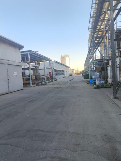 This image depicts an outdoor industrial setting, likely a factory or processing plant, located in Dimona, Israel. The scene is characterized by a clear, pale blue sky, indicating it is daytime, likely late afternoon given the warm, low-angled sunlight illuminating the structures and pipes on the right side of the frame.
The main subjects are the industrial buildings and infrastructure. On the left, a large, light-colored industrial building with a prominent door is visible. Adjacent to it is a covered walkway or lean-to structure with corrugated metal roofing. Further into the background, other industrial buildings and a tall white silo are present. Dominating the right side of the image is a complex network of elevated metal walkways and large diameter pipes, suggesting a chemical or manufacturing process.
Several objects are scattered around the yard. Pallets of blue and clear-wrapped containers, some labeled, are stacked on the ground near the pipes. There are also large green industrial bins. A red fire extinguisher is mounted on a wall near the covered walkway on the left.
The ground is paved with asphalt, showing signs of wear and cracking in places. The overall atmosphere is one of operational industry, with no people visible in the frame. The presence of the silo, the type of infrastructure, and the clear sky are typical of an industrial zone in Israel. No discernible text is legible beyond what might be labels on the containers.