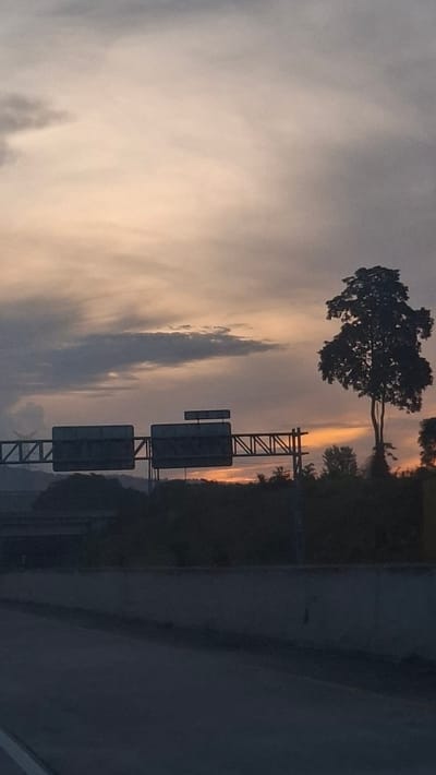 This is a photograph taken at dusk along a highway in Lonbaroh, Indonesia. The sky is overcast with muted orange and grey clouds. In the middle ground, there are two large highway signs mounted on metal gantries. To the right of the signs, a silhouetted tree stands tall against the fading light. The foreground consists of the road and a concrete barrier. The overall scene suggests a moment of transition between day and night on a major roadway, with no people or vehicles clearly visible.