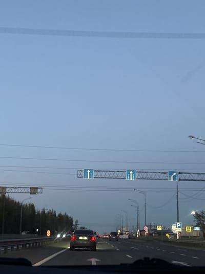 The image depicts a highway scene in Novaya Pustosh, Russia, viewed from inside a moving vehicle. The camera is oriented such that the road and adjacent areas are on the right, and the sky dominates the left. Traffic is present on the highway, with a line of cars, including a dark-colored SUV with visible taillights, indicating likely braking. Overhead, a series of traffic direction signs are mounted on gantries that span the road. These signs include directional arrows and parking symbols. Streetlights are visible along the roadside. Trees and vegetation form a barrier to the right of the highway. The sky is a muted blue, suggesting dawn or dusk.