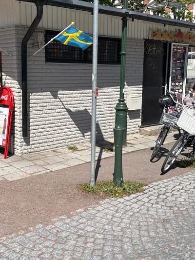 The image depicts a street scene in Malmö, Sweden, featuring a building with a Swedish flag displayed in a window. A green lamppost stands in front of the building, casting a shadow on the brick facade. A bicycle is parked near the entrance of what appears to be a small eatery or shop, indicated by the "Fast Food" sign above the door and a menu board. The sidewalk is paved with cobblestones and standard paving stones. A Coca-Cola sign is positioned near the base of the building. The scene is bathed in sunlight.