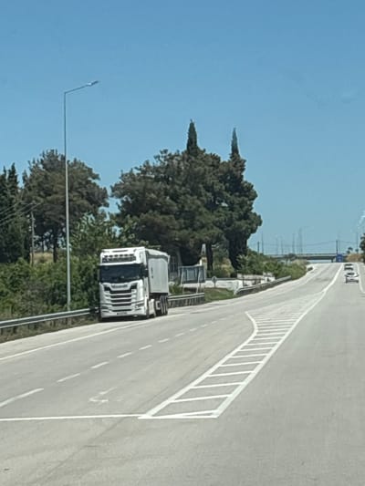 A white semi-truck with a trailer is parked on the shoulder of a highway in Lyra, Greece. The truck is facing to the left and its hazard lights are on. The highway is paved and has white lane markings. There are trees and vegetation on the left side of the highway, and a lamppost is visible. The sky is clear and blue. In the distance, a bridge can be seen over the highway, with several cars on the road.