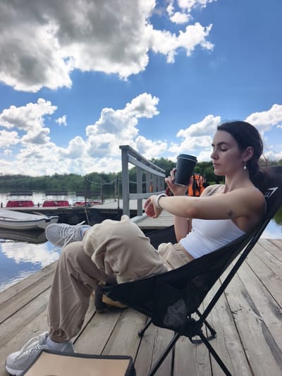 A young Caucasian woman with dark hair pulled back sits in a black portable chair on a wooden pier. She is wearing a white tank top and tan sweatpants, with white sneakers and socks. She is holding a dark-colored cup to her lips with her right hand and has a pearl earring in her left ear. A small sun tattoo is visible on her upper left arm. The pier is made of weathered wooden planks and extends over a body of water. In the background, there is a wooden railing, several red pedal boats docked, and a white boat. The far bank of the water is lined with dense green trees. The sky above is a vibrant blue with numerous fluffy white clouds. The overall setting suggests a warm, sunny day at a lakeside or riverfront location in the vicinity of Onochino, Russia.