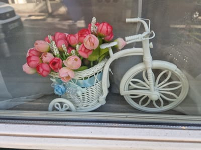 A white decorative bicycle with a basket filled with pink and red faux flowers sits on a wooden surface, visible through a window. The bicycle has a detailed front wheel with a floral pattern. The basket is adorned with a light blue ribbon and a small blue fabric flower with silver accents. Behind the bicycle, a translucent screen or fabric is visible, and beyond that, the blurred reflection of a vehicle and possibly some greenery can be seen. The window frame itself, made of a light-colored material, is in the foreground. The scene appears to be a decorative display, likely in a shop window or on a windowsill in Antony, France.