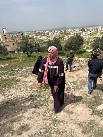 The image depicts an outdoor scene in Dura, Palestinian Territory, on a dry, uneven hillside with sparse grass and scattered olive trees. In the foreground, a woman in her 40s-50s, wearing a pink hijab and a dark purple, full-length tracksuit with white stripes, walks or stands, looking towards the viewer's left with a serious expression. Red and white striped barrier tape is visible on the ground around her feet. Behind her, to the left, a person in a black, full-length garment is partially visible from the back. To the right, a man in a dark blue jacket and jeans is seen from behind, holding a camera with a telephoto lens. Further back, another man in a dark green hooded sweatshirt and blue jeans stands, looking at a mobile phone. Several other individuals are present in the mid-ground, partially obscured. In the background, an urban settlement with numerous residential buildings, consistent with the town of Dura, spreads across a valley and up an opposing hillside. A multi-story building with a white rooftop water tank is visible on the left side of the mid-ground. A small structure made of concrete blocks is present on the ground within the area marked by the barrier tape. The sky is hazy.