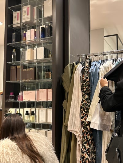 The image depicts an indoor scene within a retail store, likely a fashion and beauty outlet, specifically a Zara store, given the visible branding.
In the upper part of the frame, a person with dark long-sleeved clothing is actively browsing items on a clothing rack. Their hands are engaged in sifting through various garments, which include blue denim jeans, a black and white patterned top, a light-colored ribbed top, and other neutral-toned clothing pieces like an olive-green shirt. A piece of white paper with a circular, mandala-like design and some text is partially visible among the clothes on the rack.
Below the clothing section, a multi-tiered display shelf is filled with an array of perfume bottles and their corresponding packaging boxes. The shelves appear to be made of clear acrylic or glass, creating a neat and organized presentation for the fragrances. Many of the boxes prominently feature the brand name "ZARA," with specific variants like "ZARA WONDER ROSE" and "ZARA GARDENIA" suggested by the typical branding and color schemes (pink and brown boxes). Various perfume bottle designs are visible, ranging from clear glass to colored glass.
In the bottom right corner, the back of a second person's head and shoulders are visible. This individual has long, dark hair and is wearing a fluffy, light-colored jacket or coat, seemingly engaged in examining the perfumes on display.
The setting is well-lit with artificial indoor lighting. The activity suggests shopping or browsing merchandise. The general atmosphere is that of a modern, organized retail space.