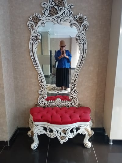 A full-length mirror with an ornate white and gold frame stands against a textured beige wall. A woman wearing a blue shirt, black skirt, and a tan hat is reflected in the mirror, holding a phone to take a picture. In front of the mirror is a tufted red bench with a carved white base and gold accents. The floor is black and reflective. This scene is set in a lobby in Kırıkkale, Turkey, likely within a hotel or administrative building.