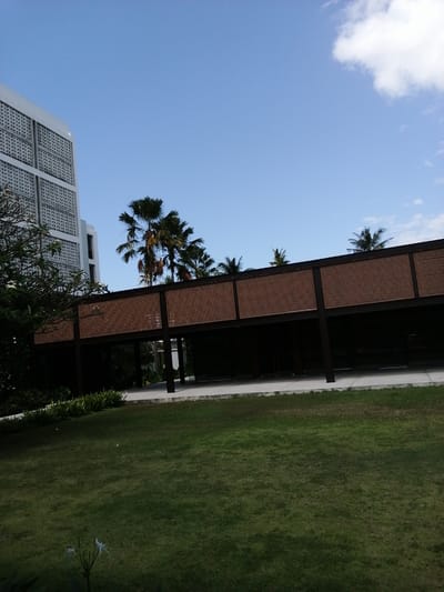 The image shows a modern building with a patterned facade and a covered walkway with a woven texture, set against a clear blue sky with a few white clouds. Palm trees are visible behind the walkway. The foreground consists of a neatly mowed green lawn with some white flowers. The scene appears to be a resort or hotel complex in Banyuwangi, Indonesia, given the architectural style and the presence of palm trees, which are common in tropical regions. There are no people or animals visible in the image, and no specific actions or interactions are depicted. The environment is clean and well-maintained, suggesting an outdoor recreational or hospitality setting.