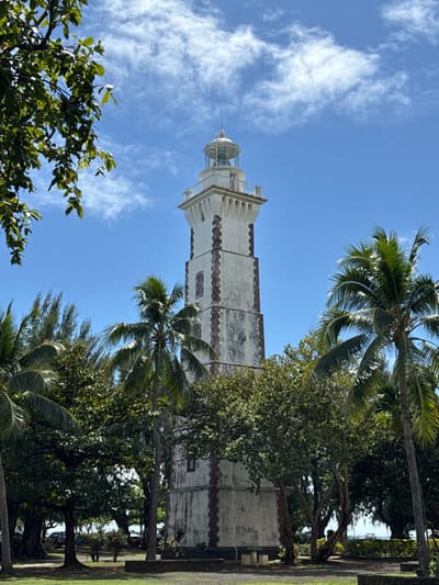 The image displays the Pointe Vénus Lighthouse in Māhina, France, under a bright blue sky with scattered white clouds. The prominent structure is a tall, square-based lighthouse, primarily white or light-colored with dark, weathered corner accents along its shaft. Multiple small, rectangular windows are visible. The upper section features a gallery with a railing, topped by a glass-enclosed lantern room with a metallic dome and finial.
The lighthouse is surrounded by dense tropical vegetation. Numerous palm trees, including tall coconut palms, are visible, some reaching the mid-section of the lighthouse. Other broad-leafed trees with lush green canopies are interspersed, with some foliage framing the upper left foreground. In the background, through breaks in the trees, a body of water is discernible, possibly indicating a coastal location. The ground in the foreground consists of grass and darker, possibly paved or graveled areas around the base of the lighthouse. No individuals or specific activities are depicted.