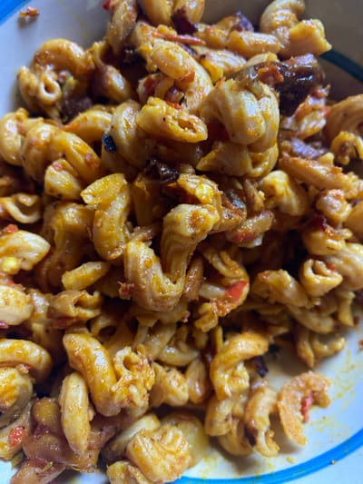 The scene depicts a close-up, overhead view of a serving of pasta in a bowl, located in Mape, Nigeria. The primary subject consists of numerous cooked cavatappi or cellentani-shaped macaroni noodles, uniformly coated in an orange-red sauce. Visible within the dish are small, irregular dark brown fragments, consistent with cooked meat or caramelized vegetables, and smaller, bright red pieces, indicative of diced peppers or tomatoes. Minute specks of yellow are also present. The pasta is contained within a white bowl, a segment of which reveals a blue rim along the bottom right edge of the frame. No individuals, actions, or specific environmental details beyond the dish are discernible.
