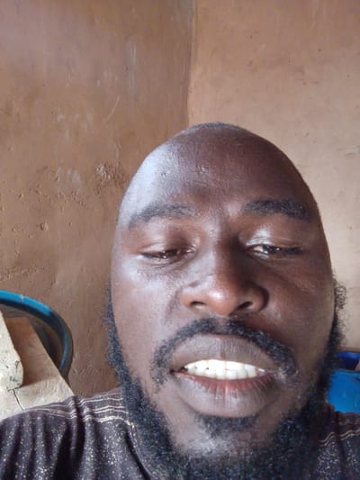 A close-up selfie shot of a dark-skinned, bald man with a full black beard and mustache, facing the camera. He is wearing a patterned shirt with light horizontal lines. His mouth is slightly open, revealing his teeth. The background consists of a textured, tan-colored wall. A blue object is partially visible to the left. There is no way to confirm the specific location (Mulke Barikin Sayejo, Nigeria) from the image alone.
