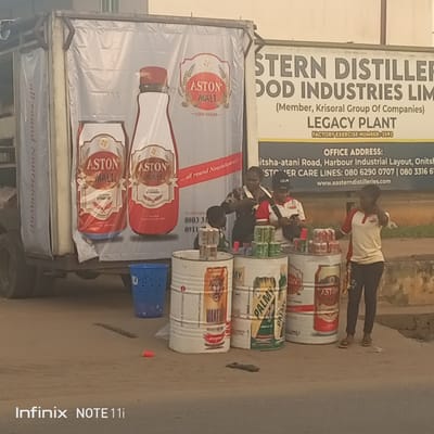 The image depicts a roadside scene in Atani, Nigeria, with street vendors selling drinks. The backdrop features a truck with an advertisement for Aston Malt and a sign for Eastern Distilleries Food Industries Limited, specifically the Legacy Plant, located on Itsha-atani Road, Harbour Industrial Layout, Onitsha. Three vendors, two men and a woman, stand behind makeshift counters made from painted oil drums displaying various beverages. A female customer is seen interacting with one of the vendors. Litter is visible on the ground. The image was taken with an Infinix NOTE 11i.