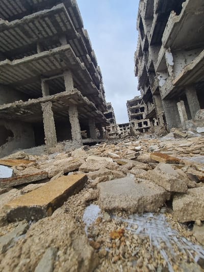 The image displays a severely damaged urban environment within Al-Qaboun Municipality, Syria, viewed from a low vantage point looking upwards. Multiple multi-story concrete buildings line a debris-filled ground, exhibiting extensive structural destruction. Their facades are largely absent, revealing exposed concrete floor slabs, columns, and internal frameworks. Many walls are disintegrated, leaving large openings and irregular, jagged edges.

The ground in the foreground and extending into the mid-distance is completely covered with a dense accumulation of rubble, consisting of fragmented concrete, broken slabs, and pulverized building materials. Beige, grey, and brown concrete pieces of varying sizes dominate the ground cover, with some discernible white, fibrous debris interspersed within the smaller gravel-like particles. No intact paving or ground surface is visible.

The sky overhead is overcast and light grey, suggesting a cloudy atmospheric condition. No individuals, animals, vehicles, or signs of recent human activity are present. The scene depicts a desolate landscape of architectural ruin and abandonment.